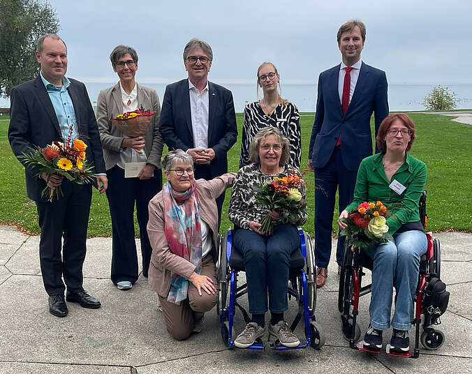 Gruppe von Männern und Frauen sowie zwei Frauen im Rollstuhl mit Blumen